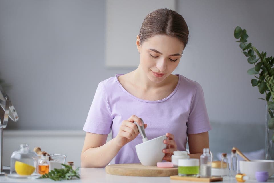 Người phụ nữ nghiền nguyên liệu trong cối sứ, trên bàn bày lọ mỹ phẩm và thảo mộc