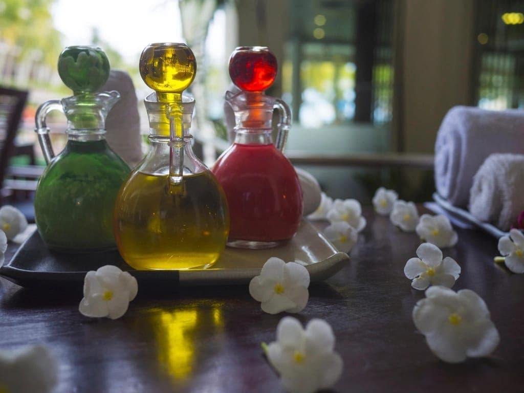 Ba bình thuỷ tinh đựng dầu xanh, vàng, đỏ trên khay, xung quanh rải hoa trắng