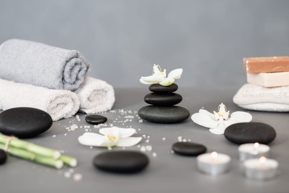 Khăn trắng cuộn tròn, đá đen xếp chồng, hoa lan trắng và nến nhỏ trên nền xám