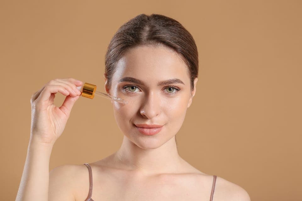 Người phụ nữ dùng ống nhỏ giọt thoa serum lên má, nền màu be