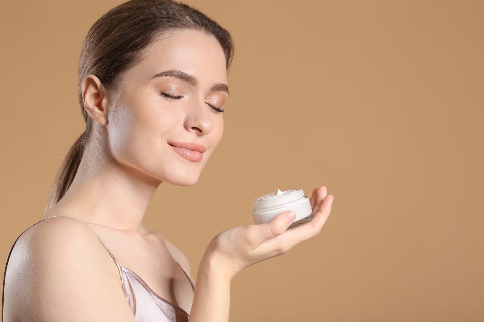 Người phụ nữ nhắm mắt, tay cầm hũ kem dưỡng mở nắp