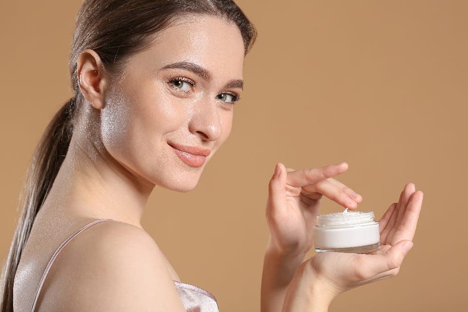 Người phụ nữ mỉm cười cầm hũ kem dưỡng đã mở nắp, ngón tay chấm kem