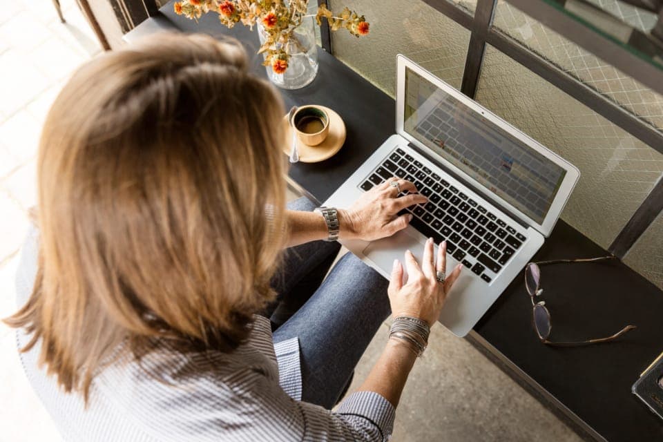 Người phụ nữ tóc ngang vai gõ laptop bên tách cà phê và bình hoa, nhìn từ phía sau