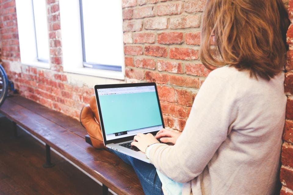 Người phụ nữ ngồi trên ghế gỗ cạnh tường gạch, gõ phím laptop đặt trên đùi